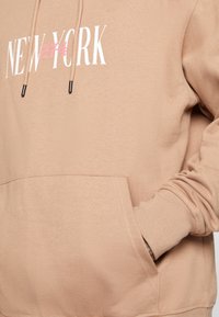 Felpa beige con tasca anteriore, cappuccio con lacci neri e grafica "NEW YORK" bianca con accentuazioni rosa. Tessuto morbido.