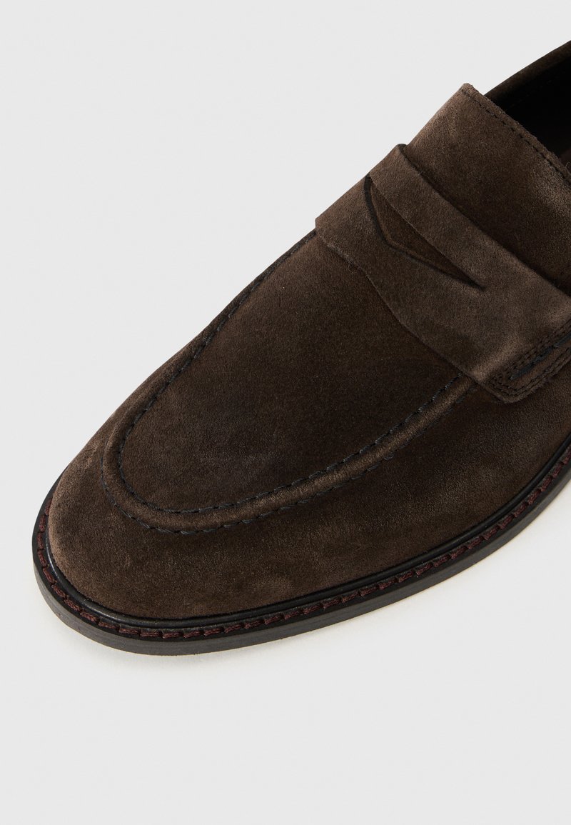 Mocassin en nubuck marron foncé avec semelle noire cousue et détail de sangle classique, présenté sur un fond gris clair.