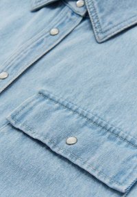 Tissu en denim bleu clair avec une poche poitrine boutonnée et des boutons-pression le long de la patte de boutonnage avant.