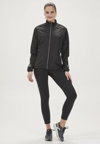 Zwarte sportjacket met een frontale rits, lange mouwen en reflecterende accenten; gecombineerd met zwarte leggings en grijze hardloopschoenen.