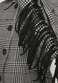 Chaqueta en blanco y negro con patrón de pata de gallo, grandes botones negros y detalles largos de flecos de cuero sintético negro en el hombro.