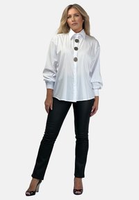 Biała koszula zapinana na guziki z trzema dużymi szarymi guzikami z przodu, z balonowymi rękawami, zestawiona z czarnymi dopasowanymi spodniami i sandałami na obcasie.