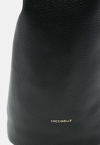 Černá kožená taška s texturovaným povrchem, s kultovním zlatým logem "COCCINELLE" na spodní části, která se vyznačuje elegantním cylindrickým designem.