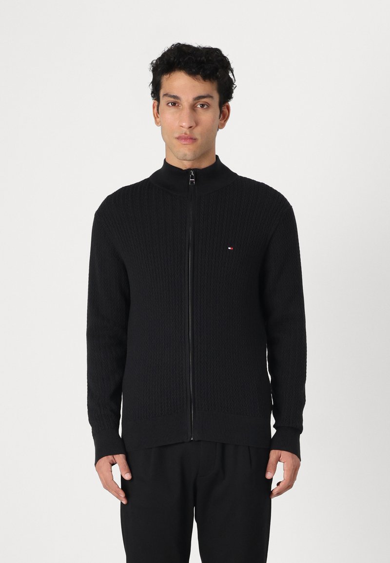 Svart stickad zip-sweater med hög krage, med ett texturerat mönster och ribbade ärmmuddar. Litet logodetalj på bröstet.