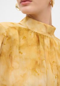 Gelbe Bluse mit hohem Kragen, die ein plissiertes Design, ein dezentes Blumenmuster und dünne vertikale Streifen aufweist. Der Stoff wirkt texturiert und leicht.
