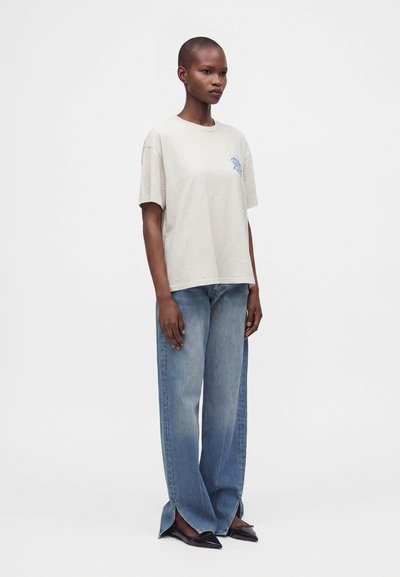 Γυναίκα που φοράει ανοιχτό μπεζ oversized μπλουζάκι, μπλε τζιν με φαρδιά μπατζάκια και εμπρόσθιες σκισίματα, και μαύρα μυτερά μπαλαρίνια, στέκεται μπροστά σε ένα απλό φόντο.