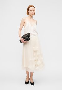 Γυναίκα με λευκό αμάνικο τοπ και μακριά φούστα από τούλι με διακοσμητικά λουλούδια, κρατώντας ένα μαύρο πλεκτό τσαντάκι και φορώντας μαύρα μυτερά παπούτσια.