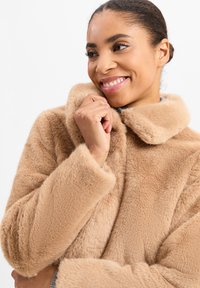 Beżowy futerko z miękką teksturą, z kołnierzem na zagięciu i szerokimi rękawami. Modelka trzymała kołnierz z lekkim uśmiechem.