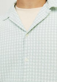 Chemise boutonnée vert clair avec un motif texturé, col court et boutons blancs, superposée à un t-shirt couleur crème.
