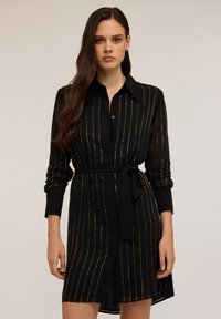 Abito nero con maniche lunghe, presenta motivi a righe verticali e una cintura da annodare. Realizzato in un materiale leggero e trasparente.