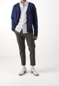 Cardigan tricoté bleu et marine avec motif en losange, chemise blanche à petits pois, pantalon en velours côtelé vert olive, et baskets blanches.