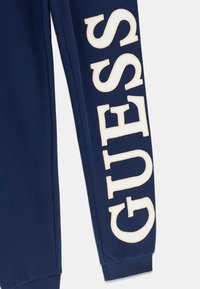 Donkerblauwe joggingsbroek met grote, geborduurde "GUESS" tekst in crèmekleur op het linkerbeen. Materiaal heeft een gladde textuur met geribde boorden.