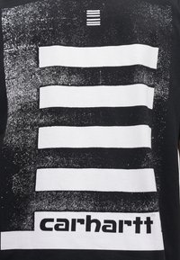 Camiseta negra con un gráfico blanco que presenta rayas horizontales desiguales y un logotipo de la marca en la sección blanca inferior. Detalle de estampado texturizado.