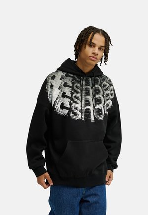 Jeune personne avec des tresses porte un sweat à capuche noir avec un grand texte graphique blanc, debout avec une main devant un jean bleu sur un fond uni.
