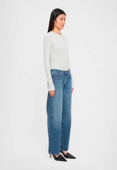 Woman with straight black hair stands sideways wearing a light gray long-sleeve top, loose blue jeans, and black pointed heels.