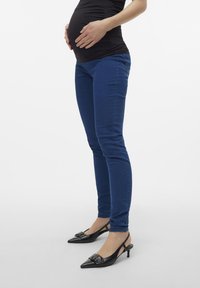 Sötétkék farmer anyás nadrág, slim fit fazonban, fekete, hegyes orrú, pánt nélküli cipővel párosítva, masnis díszítéssel.