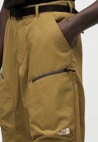 Κοντινή λήψη ατόμου που φοράει ταμπά παντελόνι τύπου cargo με το χέρι στην τσέπη, μαύρη ζώνη και λογότυπο The North Face στην τσέπη του μηρού με φερμουάρ.