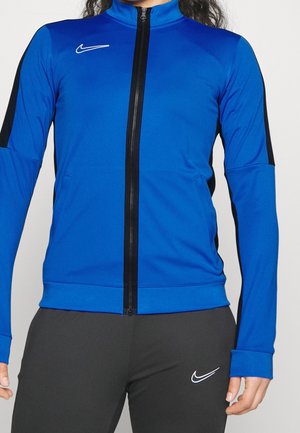 Person trägt eine blaue Nike Zip-up-Jacke mit schwarzen Seitenpaneelen und schwarze Hose mit weißem Nike-Swoosh-Logo.