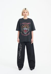 Svart oversized t-shirt med tigergrafik och texten "Ed Hardy Los Angeles", kombinerad med vida svarta byxor med silverdetaljer.