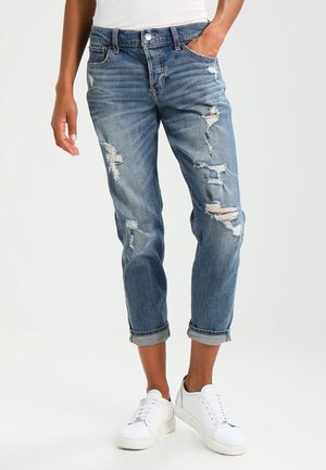 Person wearing rolled-up, ripped blue jeans and white sneakers, standing with one hand in pocket against a plain background.