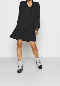 Robe mini noire à étages avec décolleté en V et manches longues bouffantes. Accents en dentelle texturée sur les manches. Portée avec des chaussettes grises et des chaussures à plateforme noires.