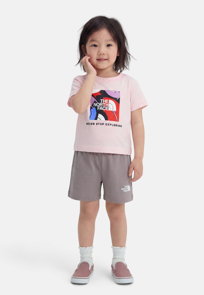 Μικρό παιδί που φοράει ροζ γραφικό μπλουζάκι North Face, γκρι σορτς, λευκές κάλτσες και ροζ παπούτσια τύπου slip-on, στέκεται με το χέρι να αγγίζει το πρόσωπο.