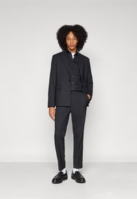 Black suit consisting of a tailored blazer and fitted trousers, paired with a black button-up shirt and black loafers, with white socks.