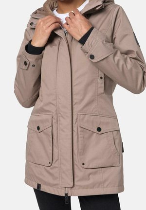 Persoon die een lichtbruine jas met capuchon draagt, voorzien van grote voorzakken en zwarte drukknopen, en de rits van de kraag verstelt.