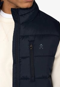 Chaleco acolchado navy con diseño de rombos, cuello alto, cremallera frontal y bolsillo en el pecho; presenta el logo encima del bolsillo y una textura lisa y brillante.
