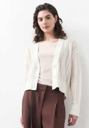 Mladá žena s kučeravými vlasmi, ktorá má na sebe biely pletený zapínací sveter cez krémové tričko a hnedé nohavice s vysokým pásom, stojaca pri bielej stene.