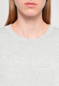 Svetlosivá mikina s okrúhlym výstrihom, na prednej strane zdobená vyrazeným textom "MICHAEL KORS", vyrobená z mäkkej, textúrovanej tkaniny.
