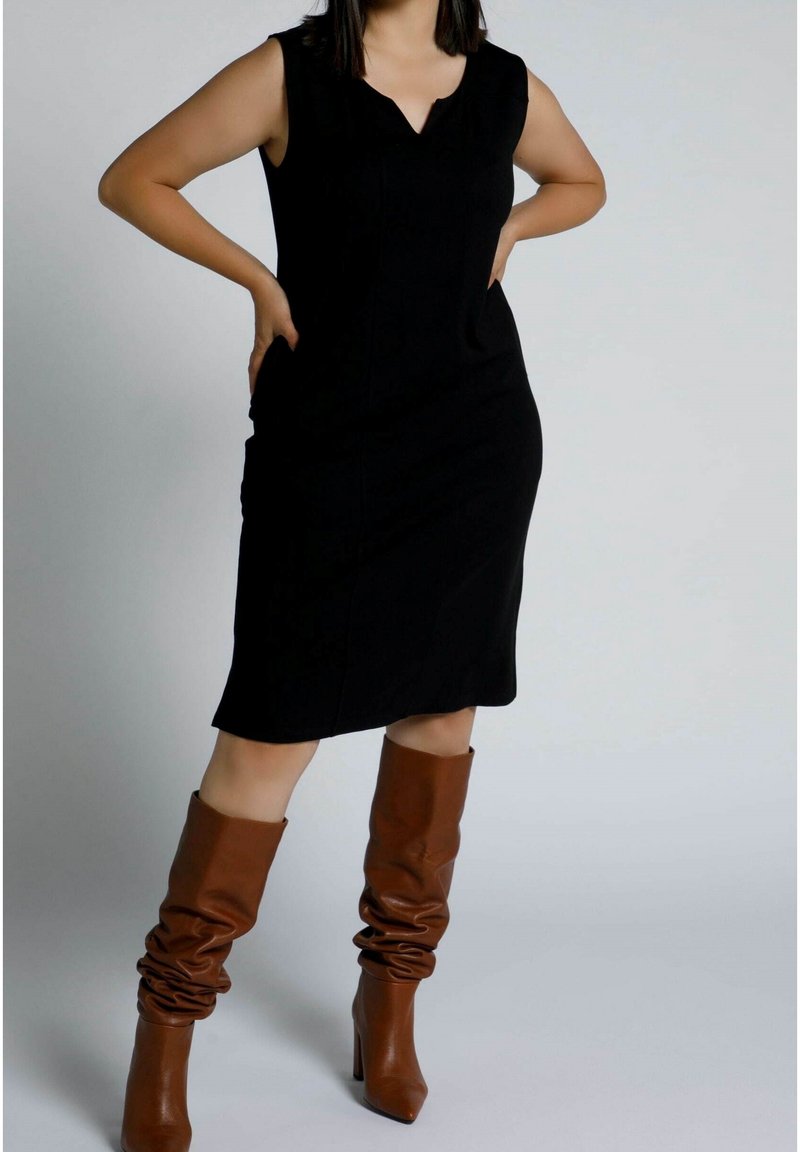 Svart ärmlös klänning med V-ringning, med en figurnära passform. Matcher med bruna knähöga, slappa klackstövlar. Slät textur.