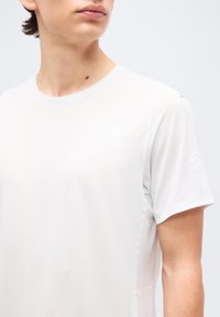 Νεαρό άτομο που φοράει απλό λευκό κοντομάνικο μπλουζάκι με στρογγυλή λαιμόκοψη, κοιτάζοντας μπροστά με το κεφάλι ελαφρώς γυρισμένο στη μία πλευρά.