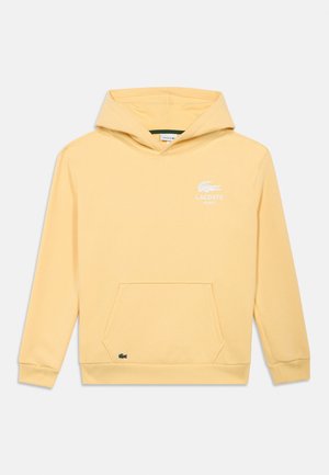 Žltá mikina s kapucňou s vreckom na prednej strane, sťahovacej šnúrky na kapucni a bielym logom Lacoste. Vyrobená z mäkkej tkaniny s rebrovanými manžetami a lemom.