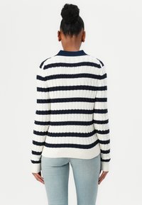 Hvit og marineblå stripete genser med ribbet tekstur og krage. Har en tettsittende silhuett og lange ermer. Bæres med lyseblå jeans.