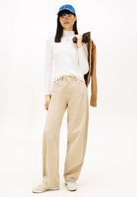 Λευκό μπλουζάκι με μακριά μανίκια και λαιμόκοψη με βολάν, μπεζ παντελόνι φαρδιάς γραμμής, καφέ σακάκι περασμένο πάνω στο χέρι και μπλε καπέλο με κείμενο. Το σύνολο ολοκληρώνουν κρεμ αθλητικά παπούτσια.