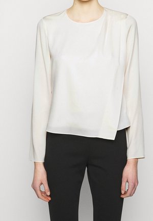 Vrouw draagt een ivoorkleurige blouse met lange mouwen en een asymmetrische zoom, gecombineerd met zwarte aansluitende broek tegen een effen achtergrond.