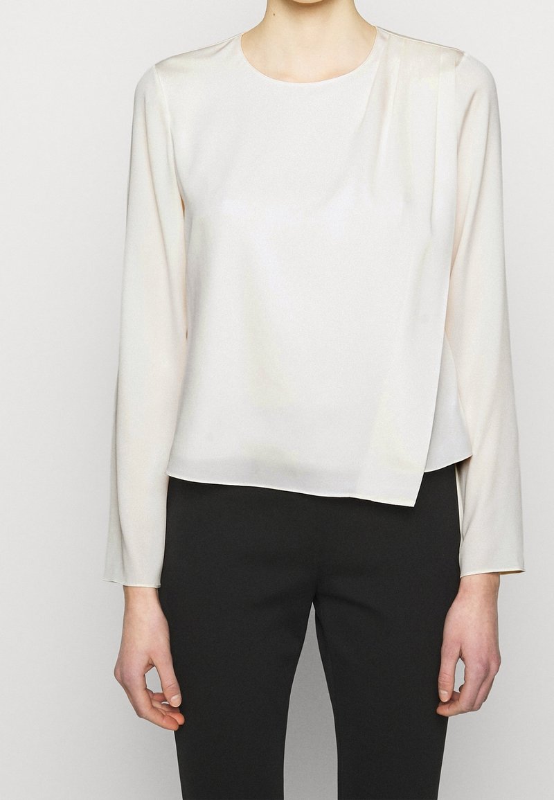 Femme portant un chemisier ivoire à manches longues avec un bas asymétrique, associé à un pantalon noir ajusté sur un fond uni.