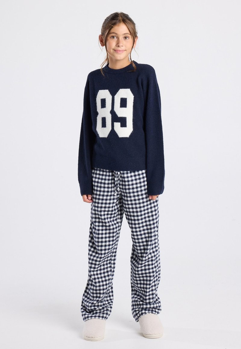 Pull marin bleu avec un design « 89 » blanc, assorti à un pantalon de pyjama noir et blanc à carreaux et des chaussures légères à enfiler.