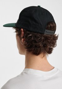 Black cotton cap with a green visor, adjustable strap, and embossed logo on the back. Curly hair visible at the nape.