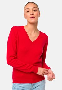 Pull rouge en V, fabriqué en maille douce avec des manches longues et des poignets côtelés, porté sur un jean bleu clair. Design minimaliste.