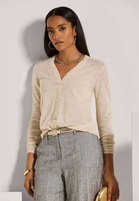 Frau in beiger, geknöpfter Strickjacke und grauen Nadelstreifenhosen, accessorisiert mit goldenen Hohrringen, einer Kettenkette und einem Armband.