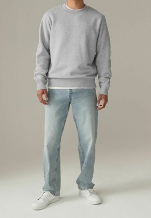 Mężczyzna w jasno szarej bluzie, jasnych niebieskich dżinsach i białych trampkach, stojący na tle jednolitego neutralnego tła.