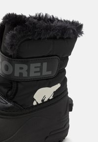 Bota negra de invierno con un suave puño de piel sintética. Presenta una correa gris etiquetada con "SOREL" y un detalle en forma de oso polar blanco en el costado.