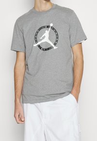 Hombre vestido con una camiseta gris de Air Jordan Flight Club y pantalones blancos, con una mano en el bolsillo y la otra colgando a un lado.