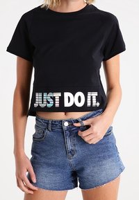 Woman wearing a black cropped t-shirt with "JUST DO IT." text and blue denim shorts, holding the waistband with one hand.