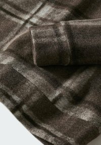 Tela de lana a cuadros marrón y gris con una textura suave, que presenta un detalle de manga enrollada y líneas sutiles que crean un patrón cuadriculado.