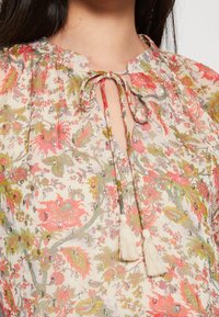 Blouse florale en tissu transparent avec des motifs colorés, dotée d'un col noué et d'accents à franges. Ornée de détails métalliques.