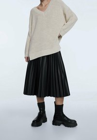 Beige gebreide trui met V-hals en geribbelde zoom, gecombineerd met een zwarte geplooide rok en zware zwarte laarzen.