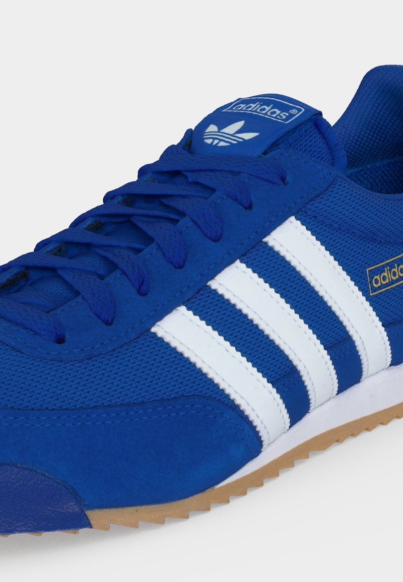 Blauwe Adidas sneaker met witte strepen, blauwe veters en een gumzool, met het Adidas-logo op de tong en zijkant.